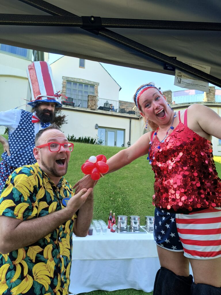 A balloon twister in a banana shirt offers a twisted balloon bracelet to a stilt walker